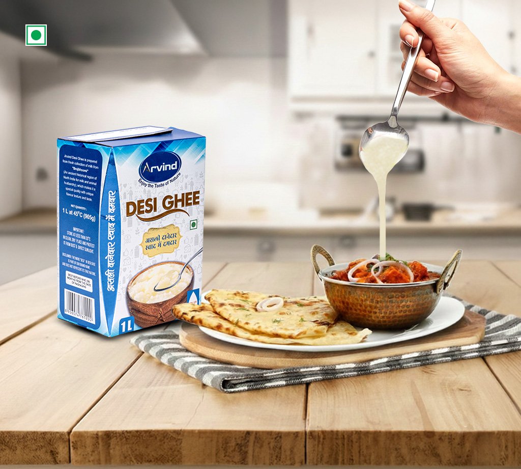 Box of Arvind Desi Ghee beside naan and curry on a wooden table as ghee is poured into the dish