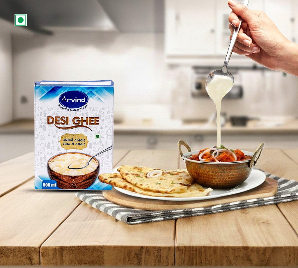 Box of Arvind Desi Ghee on a wooden table beside naan and a curry pot, with ghee being poured from a spoon into the dish