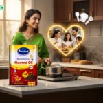 Woman in a green saree holding Arvind Kachi Ghani Mustard Oil box in a kitchen, cooking on a pot on the stove; a glowing heart frame shows a happy family dinner in the background.