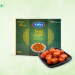 Arlys Braj Peda box with a plate of orange peda sweets in the foreground on a pale green background with a black tray nearby.