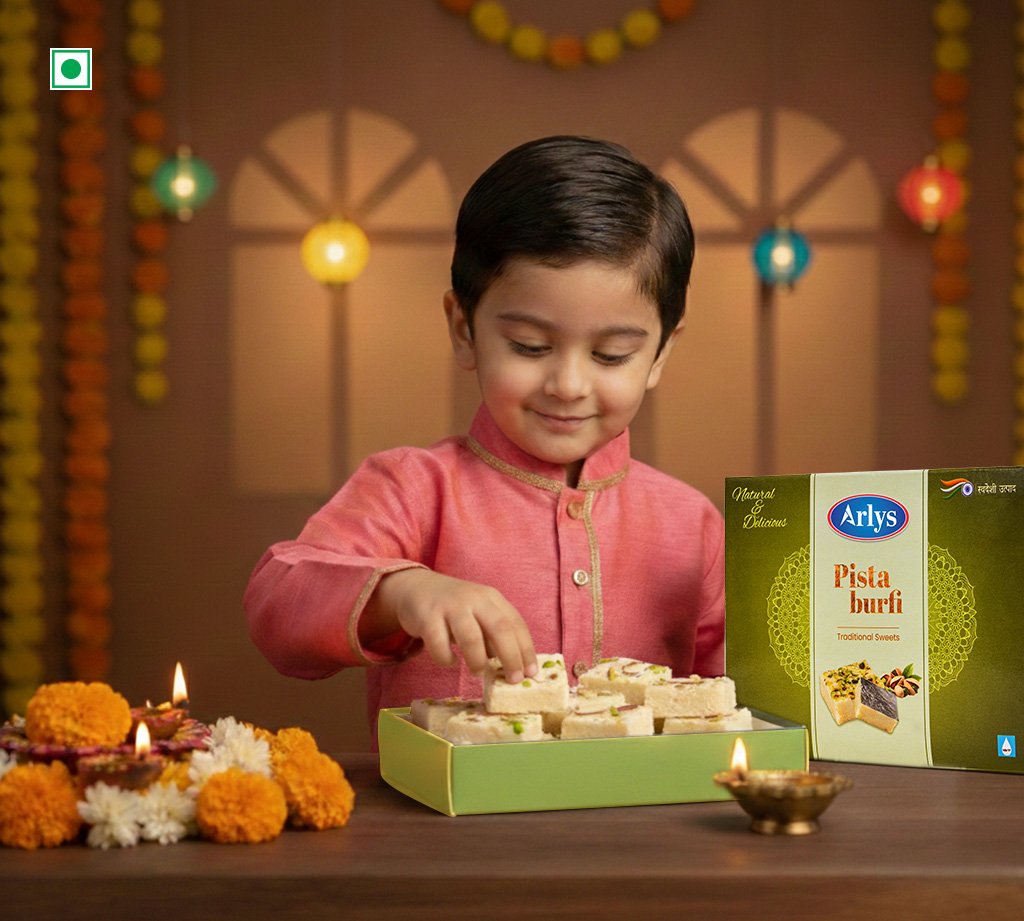 Young boy in a pink kurta selects pista burfi from a green box during a festive celebration with marigold flowers and glowing diyas on the table.
