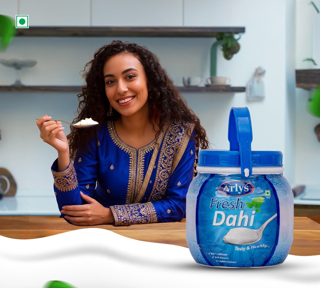Smiling woman in a blue traditional dress holds a spoonful of rice beside a large jar of Arlys Fresh Dahi yogurt on a kitchen counter.