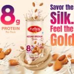 Arlys Kesar Badam Shake bottle at center with almonds and splash, promoting 8g protein per pack and gold theme chocolates and cream background