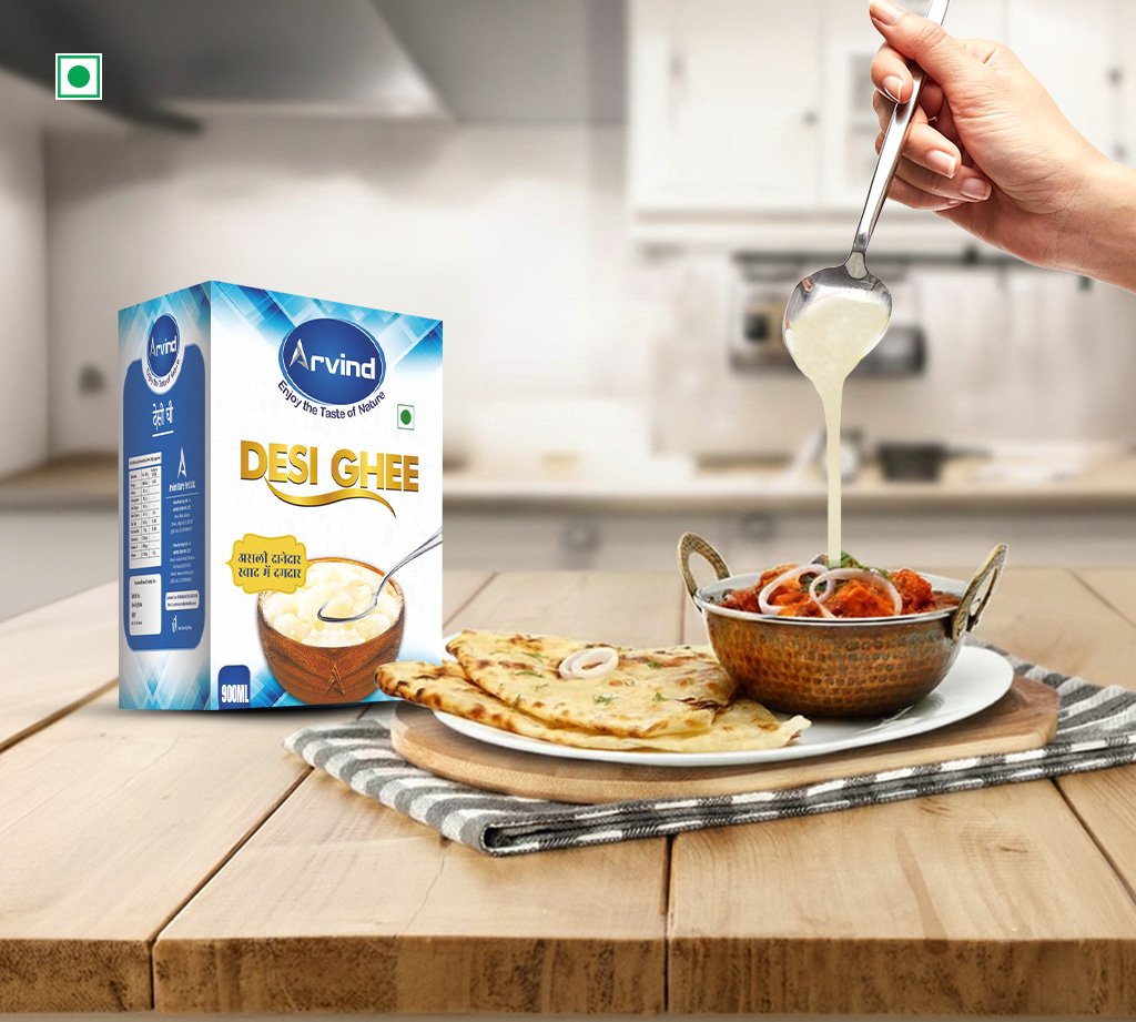 Hand pours melted ghee over a curry in a copper bowl next to naan on a wooden table, with an Arvind Desi Ghee carton in the background.