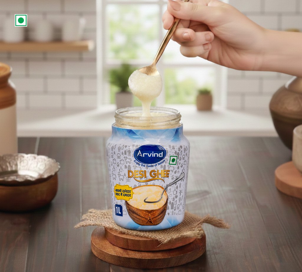 Hand holding a spoon dipping into a jar of Arvind Desi Ghee, lifting a dollop above a kitchen counter.
