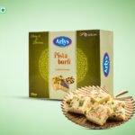Arlys Pista Burfi box with pistachio‑topped burfi pieces on a decorative plate.