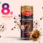 Arlys Cold Coffee cappuccino can with 8 g protein per pack, surrounded by coffee splashes and beans on a pale pink background.