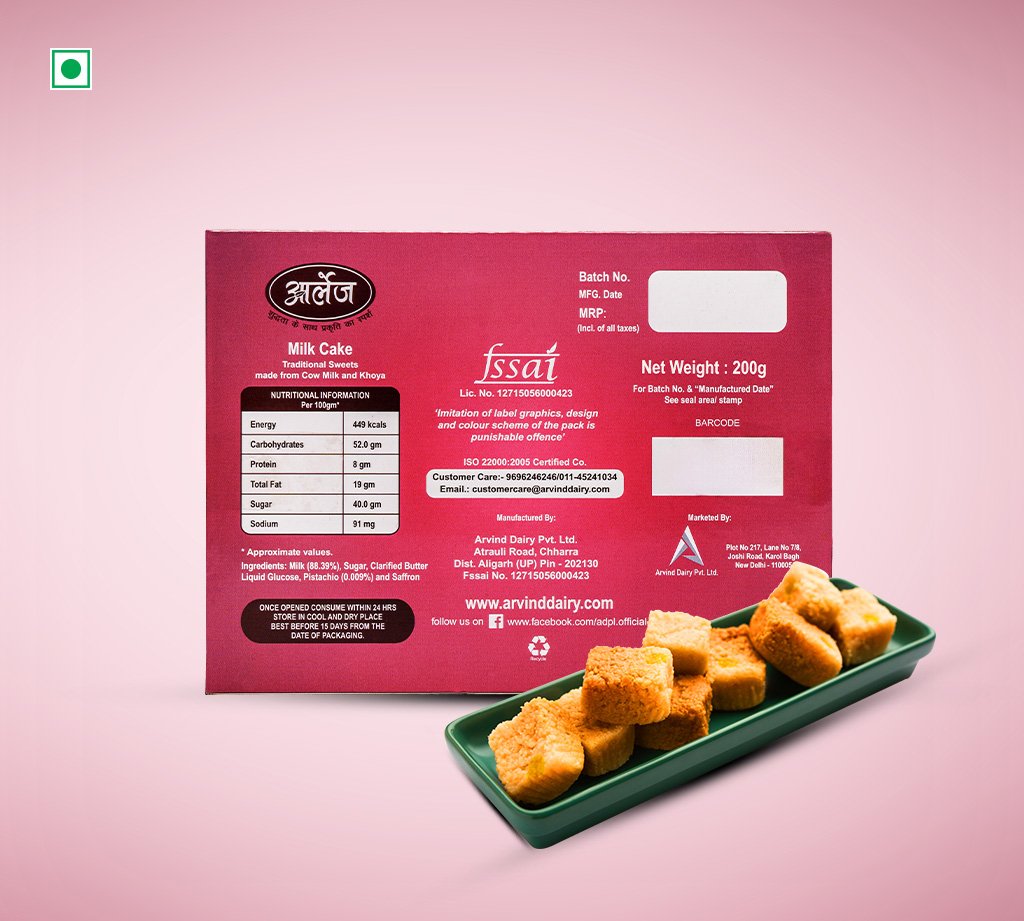 Red box labeled 'Milk Cake' with nutrition panel, batch and barcode areas, next to a green tray of cubed milk cakes on a pink background.