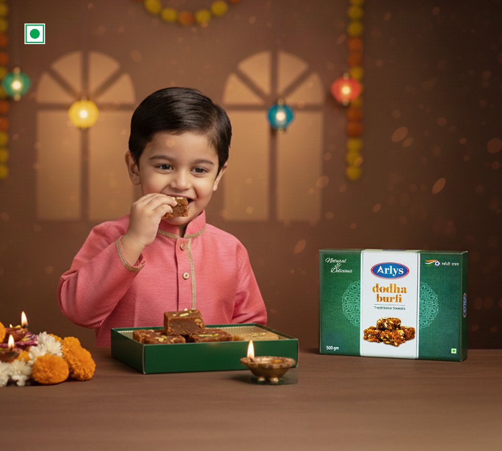 A young boy in a pink kurta eats a piece of burfi, with a green box of Arlys dodha burfi on the table beside him.