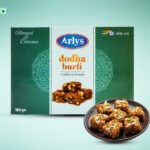 Box of Arlys dodha burfi traditional sweets, 500 g, with a plate of square burfi pieces topped with nuts.