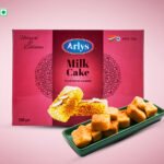 Pink Arlys Milk Cake box for traditional Sweets; plate of bite-sized fried cubes on a green tray in front.