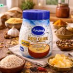 Container of Arvind Desi Ghee centered on a wooden table with bowls of spices and grains around it, in a warm kitchen setting.